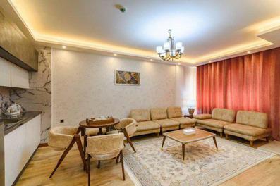 سوئیت یک خوابه هتل آرتمیس قشم با مبلمان کرم با کوسن های قرمز، لوستر، تابلو، فرش، پرده قرمز و آشپزخانه