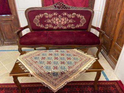 یک مبل سنتی زرشکی رنگ با پارچه مخمل که رو به روی آن یک میز قرار دارد. روی میز پارچه سنتی پهن شده است.