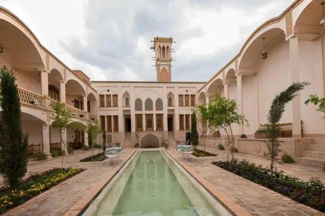 حیاط زیبای خانه محمودیه کاشان با حوض آب مرکزی، گل ها و درختان سبز، احاطه شده توسط ستون ها و اتاق های باز، یک محیط آرامشبخش و زیبا را به تصویر میکشد.