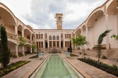 حیاط زیبای خانه محمودیه کاشان با حوض آب مرکزی، گل ها و درختان سبز، احاطه شده توسط ستون ها و اتاق های باز، یک محیط آرامش‌بخش و زیبا را به تصویر می‌کشد.