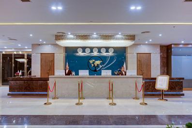 نمایی از لابی هتل خورشید هشتم مشهددو نفر در سمت چپ و راست پشت پیشخوان نشسته اند. یک لوستر بزرگ طلایی و ساعت های جهانی در بالای تصویر قرار دارند.