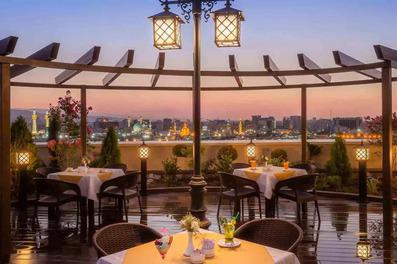 کافه رو باز هتل مدینه الجواد رو به حرم امام رضا کهمیز و صندلی های چوبی آن در تصویر دیده می شود