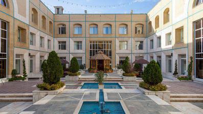 نمای بیرونی هتل قصر ضیافه مشهد با آجرهای زرد رنگ که حوض آبی رنگ و درختچه های آن در تصویر مشاهده می شود.