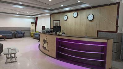 لابی هتل زاگرس با سازه ای چوبی و رنگ کرم و قهوه ای که نور بنفش روی آن تابانده شده. ساعت های جهانی بالاس سر کانتر لابی روی دیوار با رنگ قهوه ای روشن قرار گرفته اند.