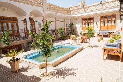 نمایی از گوشه یک حیاط قدیمی سنتی به سبک ایرانی که حوض در وسط و تخت های سنتی و گلدان در اطراف آن قرار دارد.